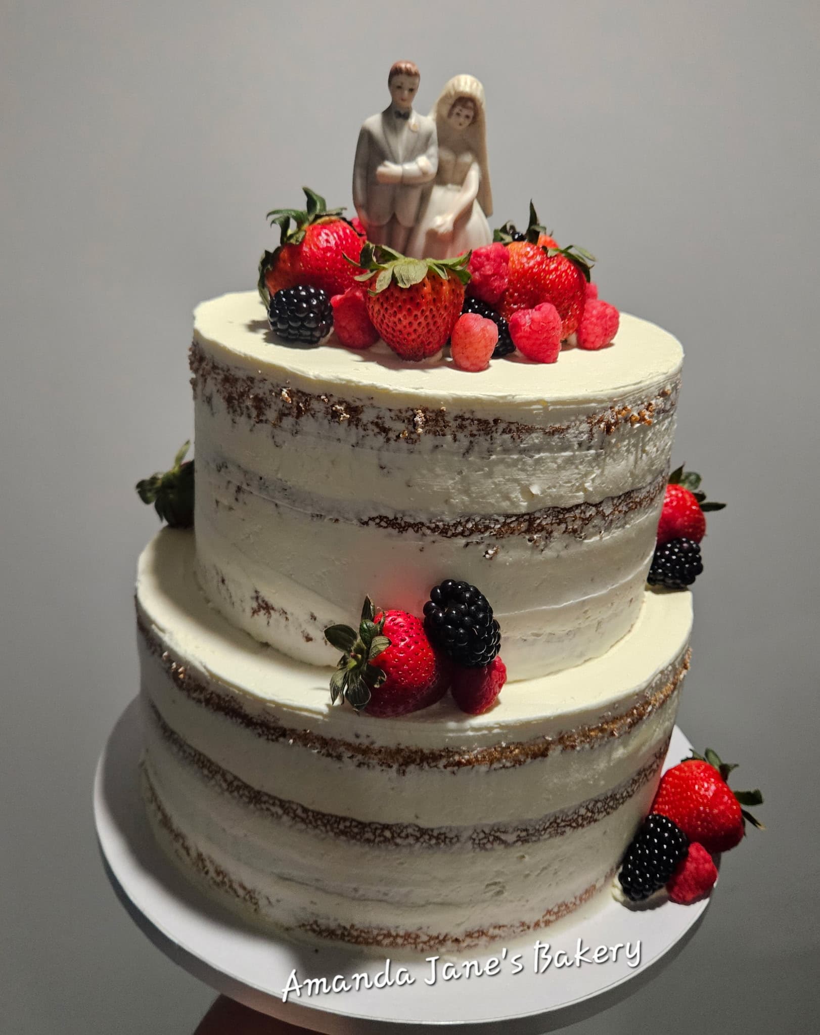 Two-tier semi-naked wedding cake with fresh berries and a bride and groom topper